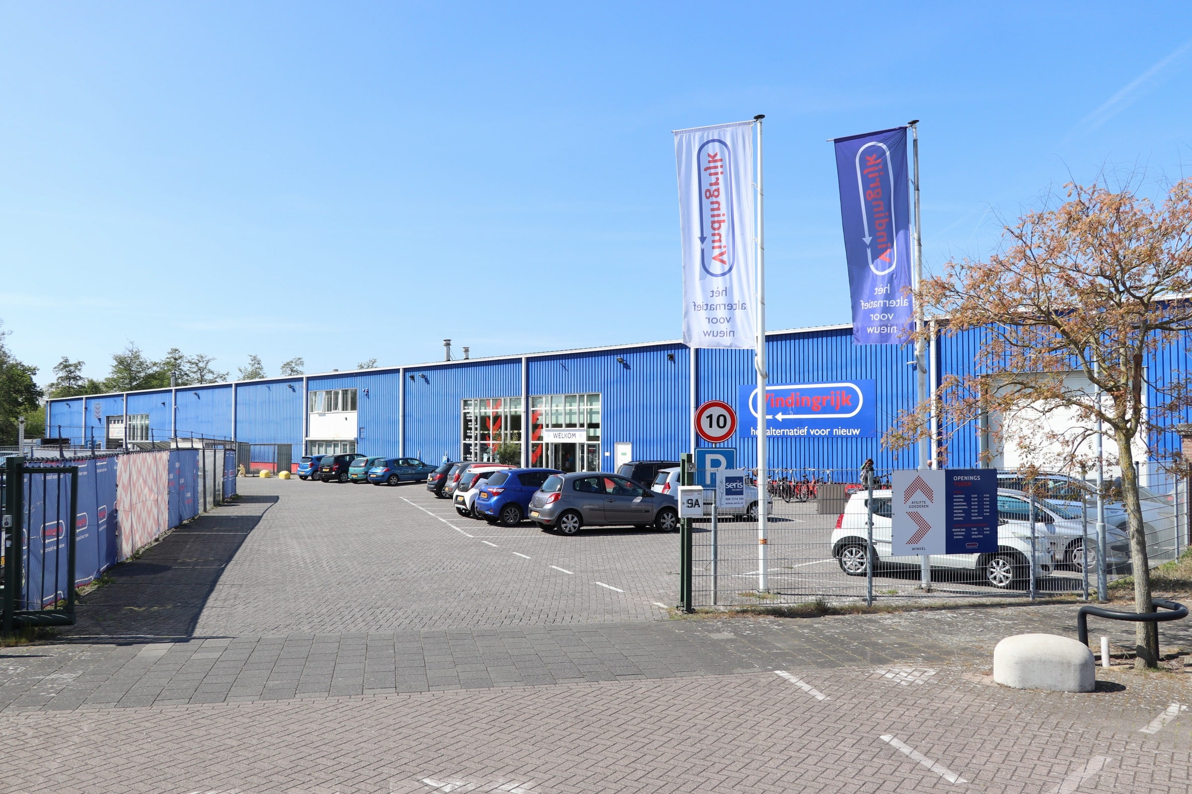 Op de foto zie je de voorkant van onze winkel. Op de parkeerplaats staan auto's geparkeerd. Rechts staan er twee vaandels met het logo van Vindingrijk.