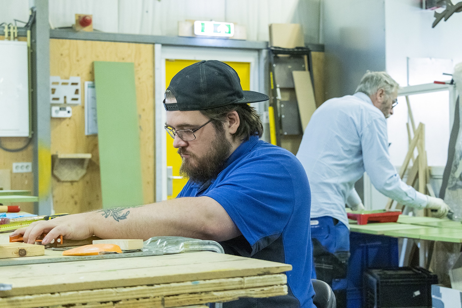 Op de foto zie je twee van onze medewerkers in de meubelwerkplaats. Op de voorgrond werkt onze collega met hout. Op de achtergrond zie je onze collega schilderen.