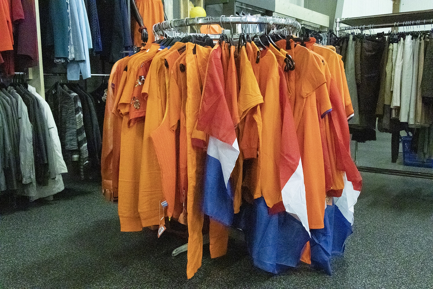 Op de foto zie je een ronde kledingrek in onze winkel. Aan dat kledingrek hangen verschillende oranjekledingstukken. Een aantal van deze kledingstukken heeft de Nederlandse vlag.